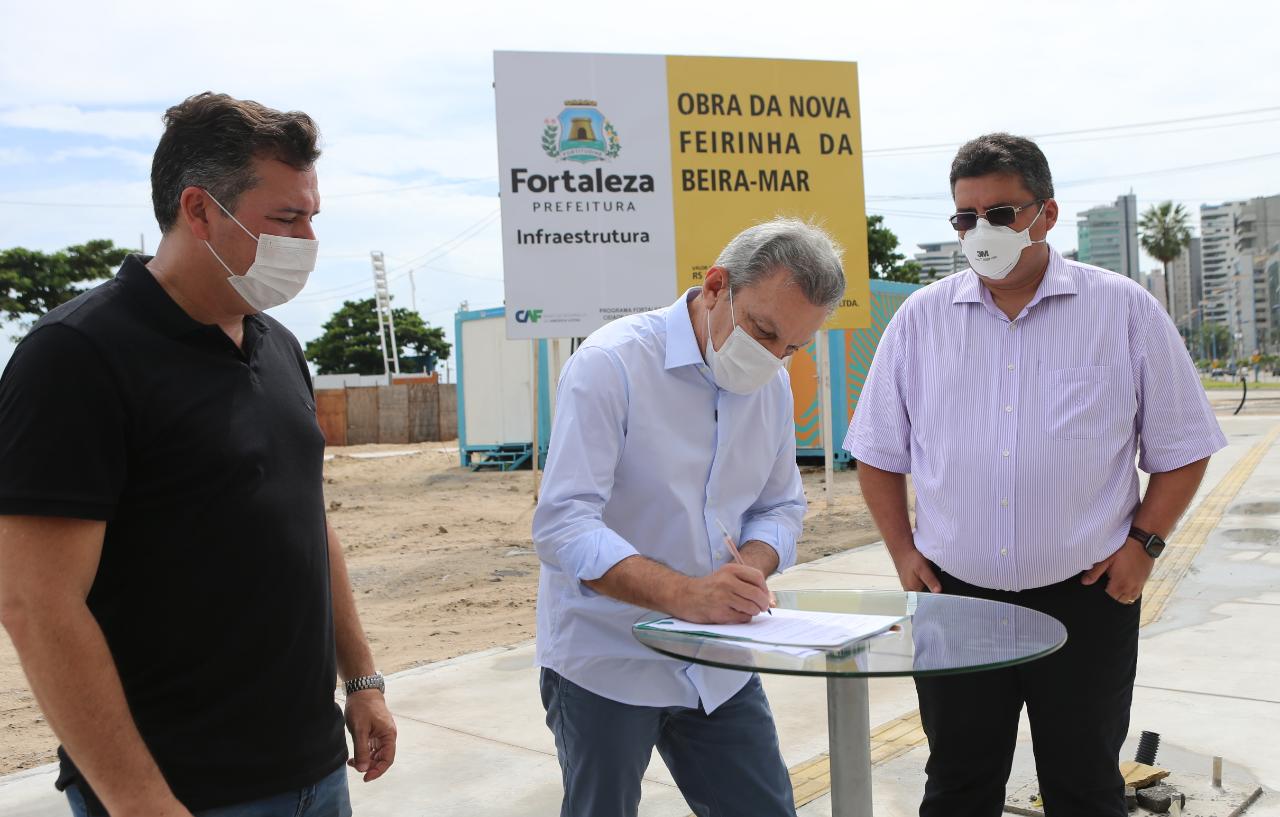 Sarto assina a ordem de serviço e, ao fundo, se vê a placa com o nome: obra da nova feirinha da beira-mar